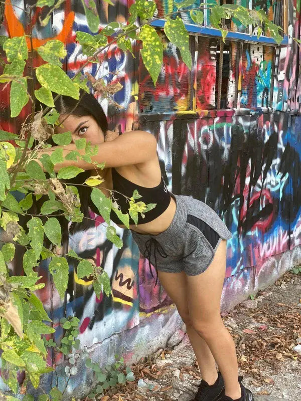 Person partially hidden behind leafy plants next to a graffiti-covered wall.