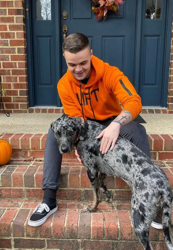 Person in an orange sweatshirt petting a spotted dog on brick steps.