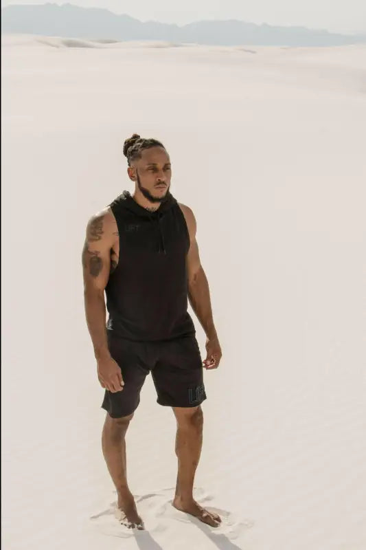 Man with tattoos wearing a black tank top and shorts standing barefoot.