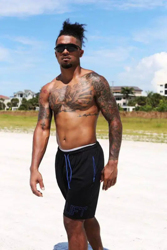 Shirtless man with tattoos and sunglasses wearing black shorts.