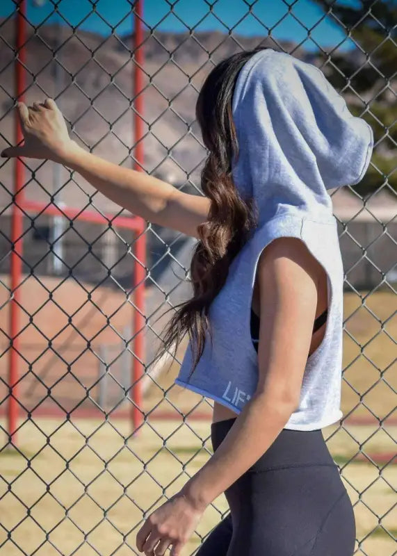 Person wearing a light blue hooded sleeveless top and dark pants, standing by a chain-link fence.
