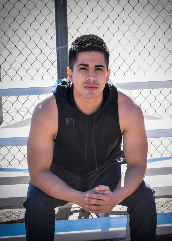 Young man with short dark hair wearing a black sleeveless shirt, sitting on a bench.