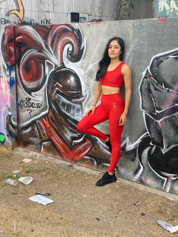 Woman in a red crop top and matching leggings posing against a graffitied wall.