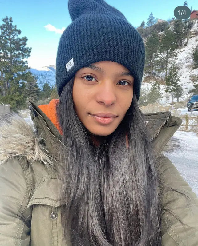 Person wearing a navy blue knit beanie and olive green winter coat in a snowy outdoor setting.